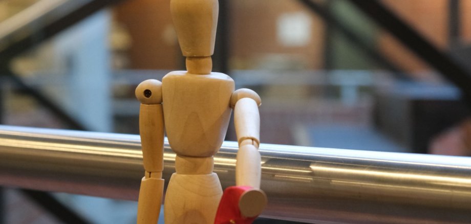 Ein Männchen aus Holz hält eine rote Schleife in der Hand, davor ein Kugelschreiber der Kreisverwaltung SÜW, ein Kondom und ein Plüschbär der AIDS-Hilfe.