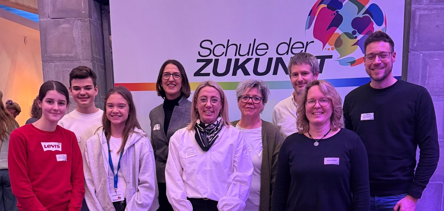 Gruppenbild: Schülerinnen, Schüler und Lehrkräfte der Realschule plus im PAMINA-Schulzentrum Herxheim mit Schulleiterin Magdalena Mayer. Im Hintergrund ist ein Rollup mit der Aufschrift "Schule der Zukunft" zu sehen.