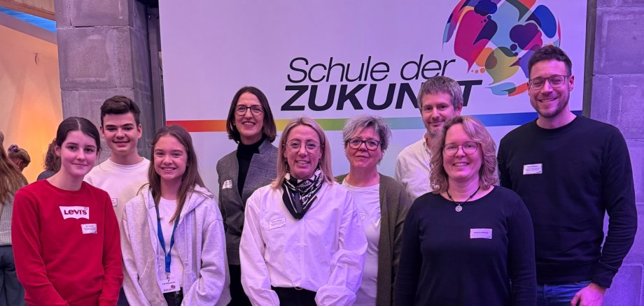 Gruppenbild: Schülerinnen, Schüler und Lehrkräfte der Realschule plus im PAMINA-Schulzentrum Herxheim mit Schulleiterin Magdalena Mayer. Im Hintergrund ist ein Rollup mit der Aufschrift "Schule der Zukunft" zu sehen.
