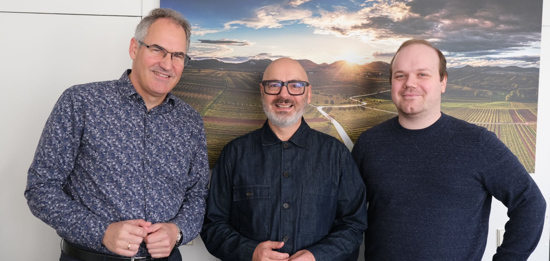 Landrat Dietmar Seefeldt, MRN-Kulturbüro-Leiter Robert Montoto und Projektleiter Sebastian Mauß vor einer weißen Wand im Kreishaus der Südlichen Weinstraße. Im Hintergrund hängt ein Bild einer Weinbergslandschaft im Sonnenuntergang.