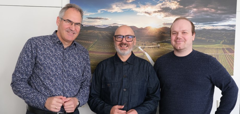 Landrat Dietmar Seefeldt, MRN-Kulturbüro-Leiter Robert Montoto und Projektleiter Sebastian Mauß vor einer weißen Wand im Kreishaus der Südlichen Weinstraße. Im Hintergrund hängt ein Bild einer Weinbergslandschaft im Sonnenuntergang.