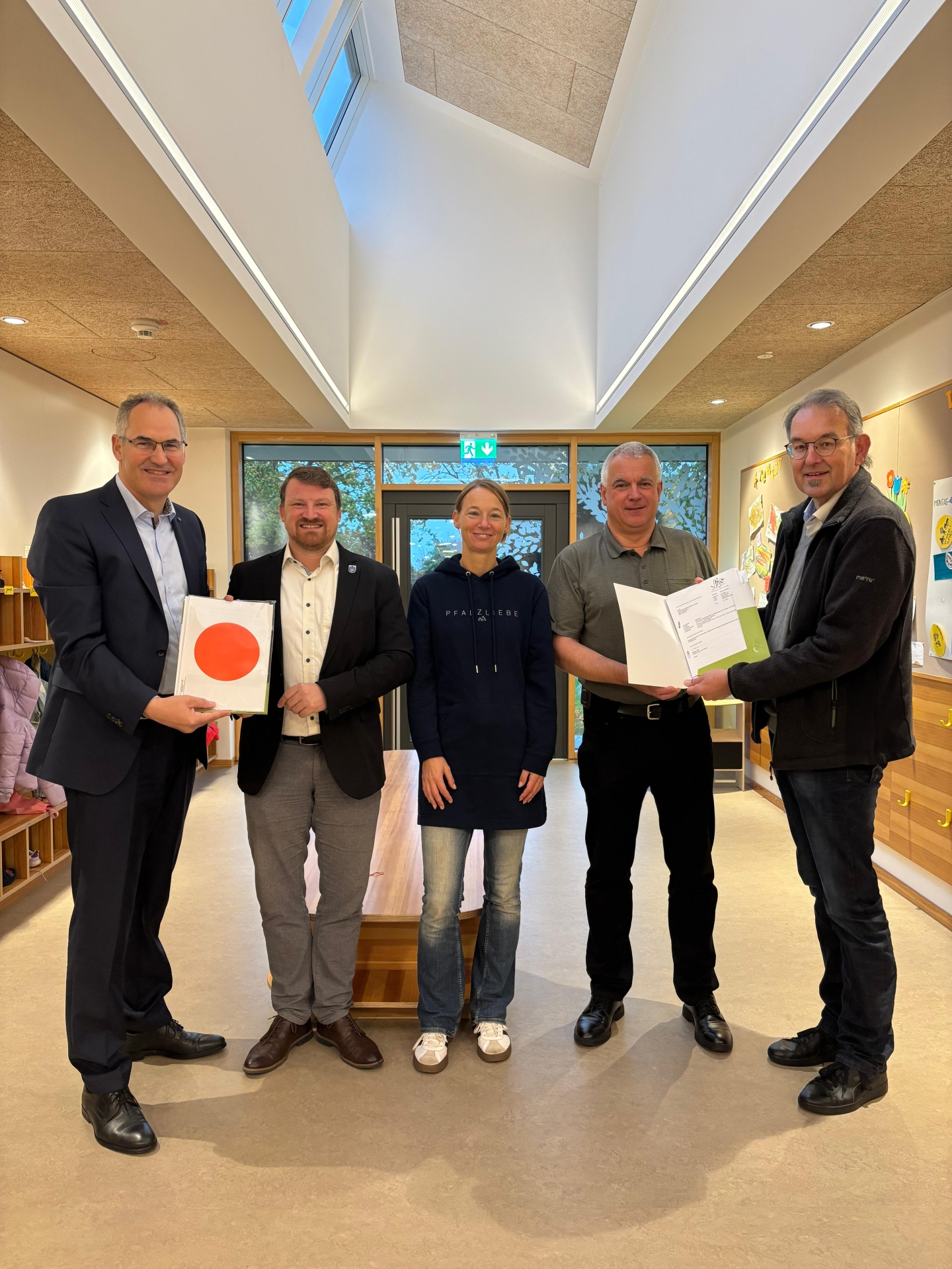 Gruppenfoto bei der Übergabe der Baugenehmigung für die kommunale Kindertagesstätte in Herxheim: Fünf Personen stehen in der hellen Eingangshalle der Kita und halten die Unterlagen der Genehmigung in den Händen.