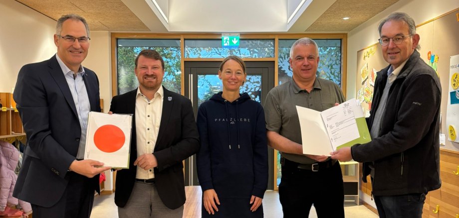 Gruppenfoto bei der Übergabe der Baugenehmigung für die kommunale Kindertagesstätte in Herxheim: Fünf Personen stehen in der hellen Eingangshalle der Kita und halten die Unterlagen der Genehmigung in den Händen.