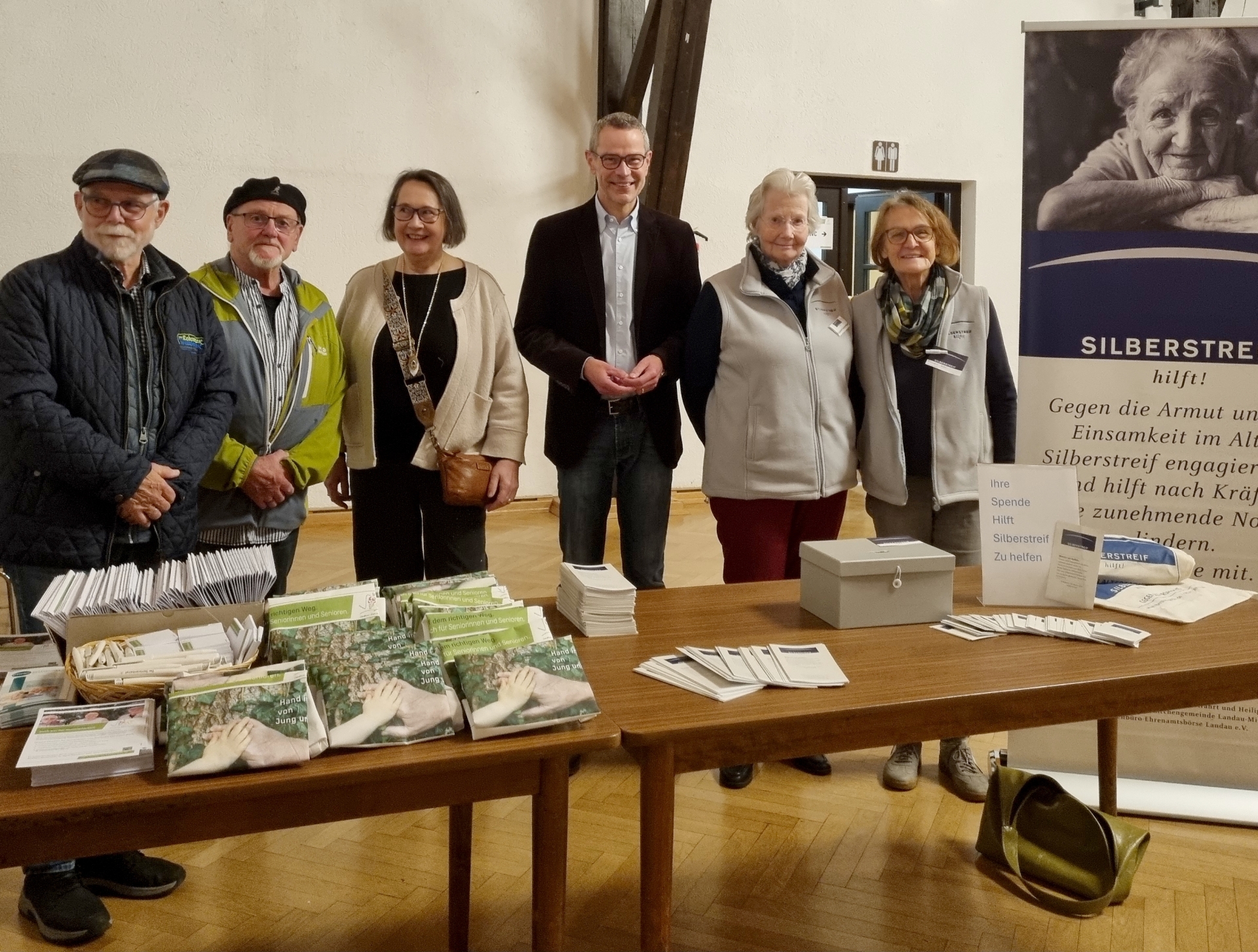 Sechs Personen stehen in einem Raum hinter zwei Tischen mit zahlreichen Broschüren und Infomaterial für ein Gruppenbild zusammen.