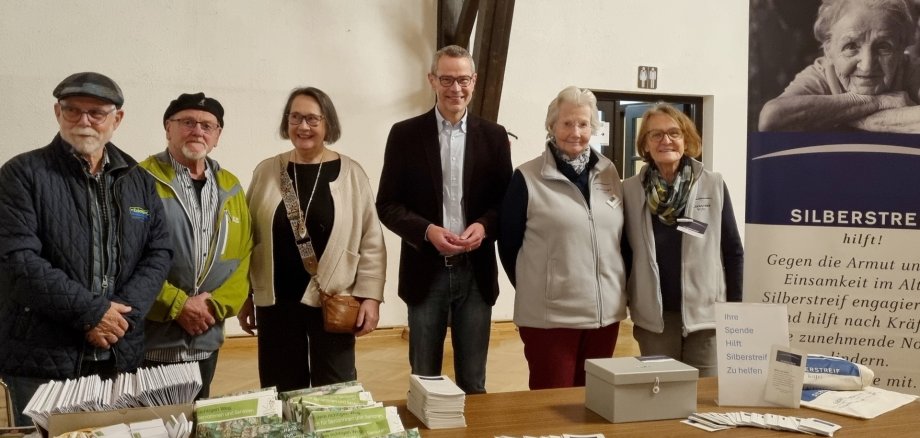 Sechs Personen stehen in einem Raum hinter zwei Tischen mit zahlreichen Broschüren und Infomaterial für ein Gruppenbild zusammen.