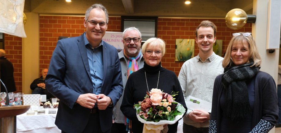 Eine Gruppe von fünf Personen steht in einem hellen Innenraum. Eine Frau in der Mitte hält einen Blumenstrauß, die anderen Personen lächeln in die Kamera. Im Hintergrund sind Tische mit Ausstellungsstücken und Kunstwerken zu sehen.