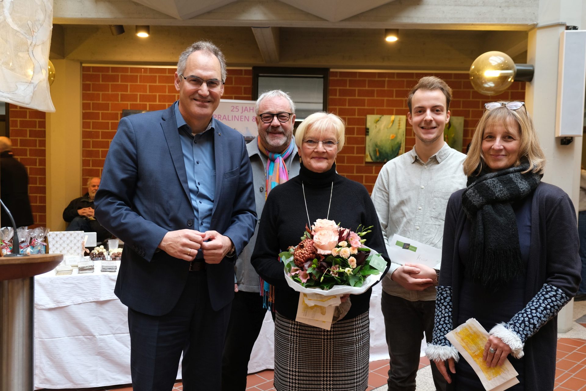Eine Gruppe von fünf Personen steht in einem hellen Innenraum. Eine Frau in der Mitte hält einen Blumenstrauß, die anderen Personen lächeln in die Kamera. Im Hintergrund sind Tische mit Ausstellungsstücken und Kunstwerken zu sehen.