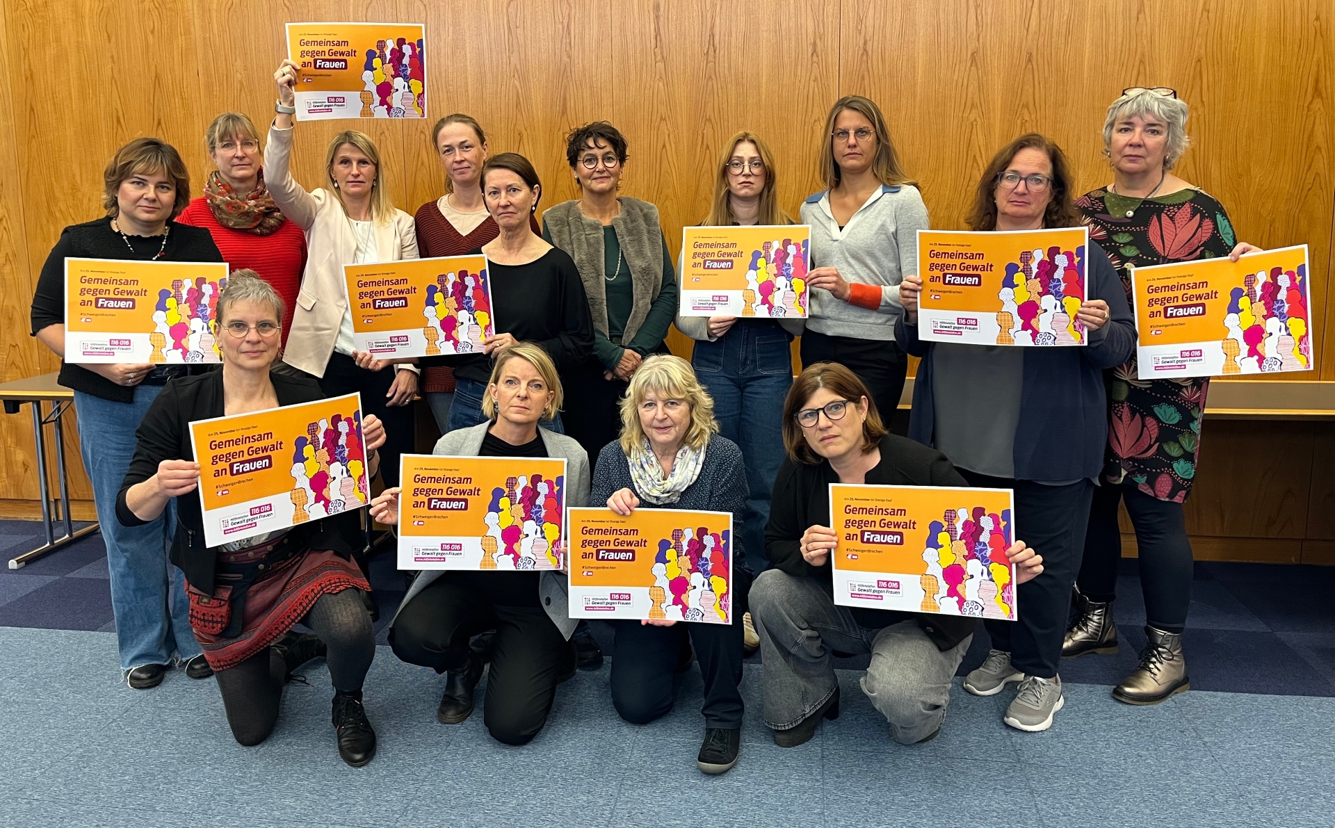 Eine Gruppe von Frauen hält Plakate mit der Aufschrift „Gemeinsam gegen Gewalt an Frauen“ in die Kamera. Die Gruppe steht in einem Raum mit Holzwand.