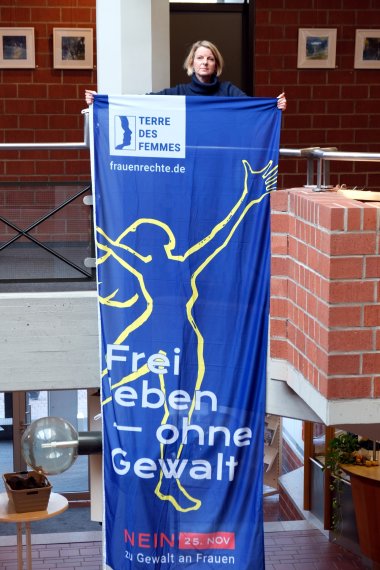 Die SÜW-Gleichstellungsbeauftragte Isabelle Stähle zeigt die Flagge, die sie am 25. November gemeinsam mit Landrat Dietmar Seefeldt, Kathrin Flory, Bürgermeisterin der Verbandsgemeinde Bad Bergzabern, und der VG-Gleichstellungsbeauftragten Marita Rothmann vor der VG-Verwaltung in Bad Bergzabern hissen wird. Foto: KV SÜW Eine Frau steht in einem Gebäude und hält eine große blaue Fahne mit der Aufschrift „Frei leben – ohne Gewalt“ und dem Logo von Terre des Femmes.