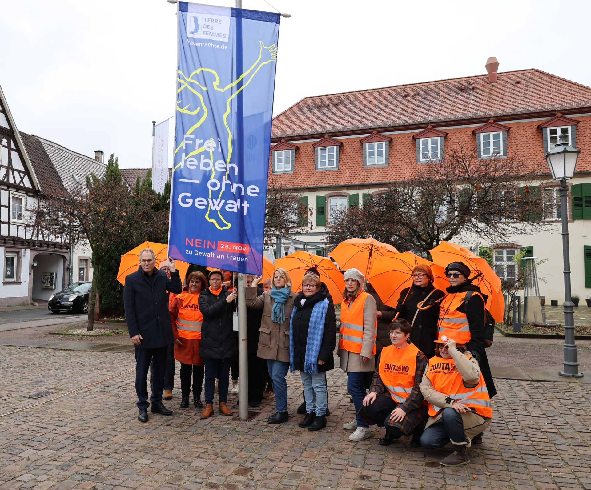 Eine Gruppe von Menschen steht vor dem Schloss Bad Bergzabern und hält eine große blaue Fahne mit der Aufschrift „Frei leben – ohne Gewalt“. Einige Teilnehmende tragen orangefarbene Westen und halten orangefarbene Regenschirme.
