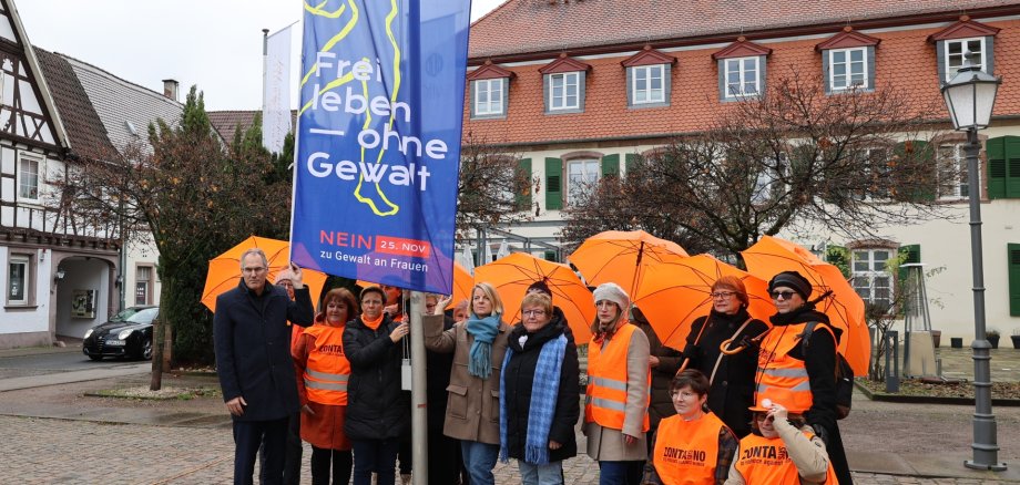 Eine Gruppe von Menschen steht vor dem Schloss Bad Bergzabern und hält eine große blaue Fahne mit der Aufschrift „Frei leben – ohne Gewalt“. Einige Teilnehmende tragen orangefarbene Westen und halten orangefarbene Regenschirme.