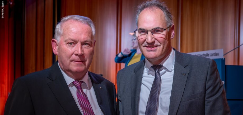 Udo Vogel (links) mit Landrat Dietmar Seefeldt: Neues Projekt „Mensa Plus“ in Bad Bergzabern steht in den Startlöchern. Foto: Rolf H. Epple Zwei Männer in Anzügen stehen nebeneinander und blicken in die Kamera. Im Hintergrund ist eine Bühne mit einer Rednerin zu erkennen.