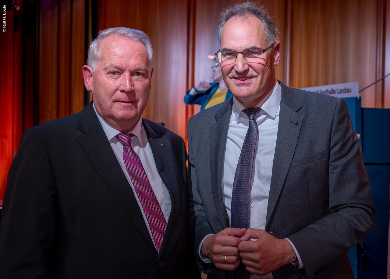 Zwei Männer in Anzügen stehen nebeneinander und blicken in die Kamera. Im Hintergrund ist eine Bühne mit einer Rednerin zu erkennen.