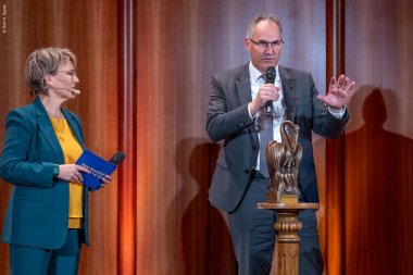 Landrat Dietmar Seefeldt gab bei der Hans-Rosenthal-Gala bekannt, dass die Dieter-Kissel-Stiftung und der Buchmann-Stiftung das warme Mittagessen am Bergzaberner Gymnasium möglich machen. Foto: Rolf H. Epple Ein Mann und eine Frau stehen auf einer Bühne mit Holzverkleidung. Die Frau hält ein Mikrofon und Moderationskarten, der Mann spricht in ein Mikrofon und steht neben einer Bronzeskulptur auf einem Podest.
