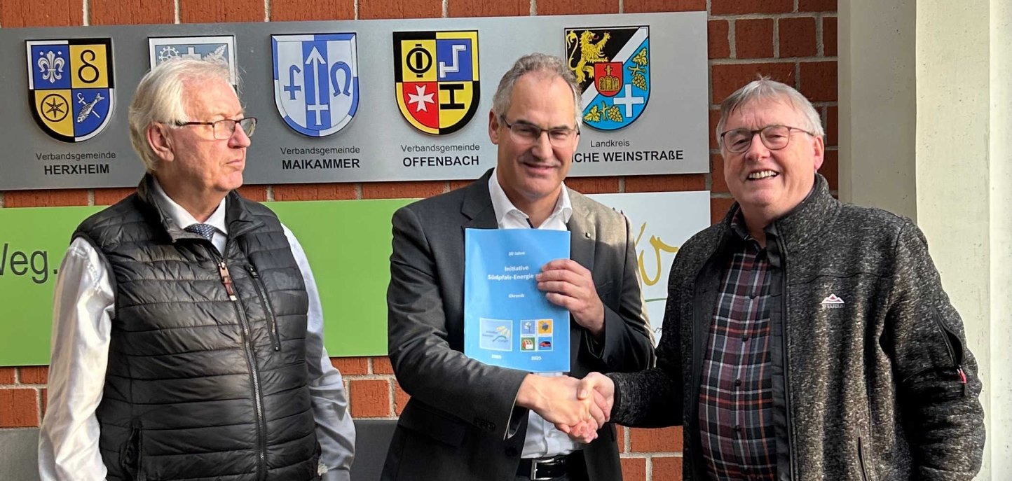 Prof. Dr. Karl Keilen, Landrat Dietmar Seefeldt und ISE-Vorsitzender Wolfgang Thiel, der die Chronik übergab stehen zusammen vor einer Ziegelwand im Kreishaus der Südlichen Weinstraße.