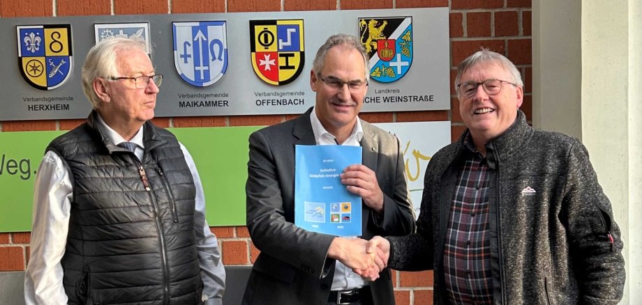 Prof. Dr. Karl Keilen, Landrat Dietmar Seefeldt und ISE-Vorsitzender Wolfgang Thiel, der die Chronik übergab stehen zusammen vor einer Ziegelwand im Kreishaus der Südlichen Weinstraße.