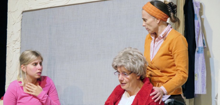 Szene aus dem Theaterstück „Maria hilf“. Foto: Helmut Dudenhöffer Drei Personen stehen auf einer Theaterbühne: Eine Frau in pinker Kleidung sitzt links und wirkt überrascht, eine ältere Frau in einem roten Bademantel sitzt im Rollstuhl in der Mitte, daneben steht eine weitere Frau in orangefarbener Kleidung und legt beruhigend die Hand auf ihre Schulter.