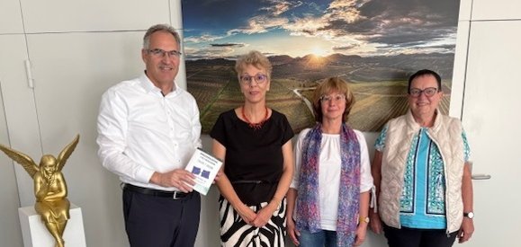 Dietmar Seefeldt, Irene Schäfer, Dr. Heike Greczko und Margret Meier-Wohlwend (von links). Foto: KV SÜW Ein Mann und drei Frauen stehen für ein Gruppenbild vor einer weißen Wand in einem Büroraum. Hinter ihnen ist ein großes Bild einer Weinbergslandschaft im Sonnenuntergang zu sehen. Am linken Bildrand ist eine kleine goldfarbene Statuette eines Engels zu sehen, der auf einem kleinen weißen Deko-Pfeiler sitzt.