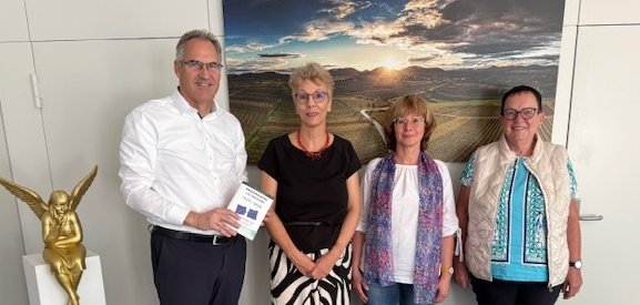 Ein Mann und drei Frauen stehen für ein Gruppenbild vor einer weißen Wand in einem Büroraum. Hinter ihnen ist ein großes Bild einer Weinbergslandschaft im Sonnenuntergang zu sehen. Am linken Bildrand ist eine kleine goldfarbene Statuette eines Engels zu sehen, der auf einem kleinen weißen Deko-Pfeiler sitzt.