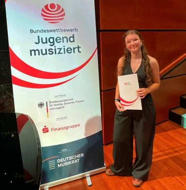 Eine junge Frau steht neben einem Rollup mit der Aufschrift "Bundeswettbewerb Jugend musiziert. Sie hält eine Urkunde in der Hand und blickt in die Kamera.