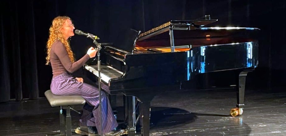 Eine junge Frau sitzt auf einer Bühne an einem schwarzen Flügel und singt in ein Mikrofon. Sie trägt ein dunkelbraunes Oberteil und eine lila Hose. Die Szene ist in warmes Bühnenlicht getaucht, der Hintergrund ist dunkel.