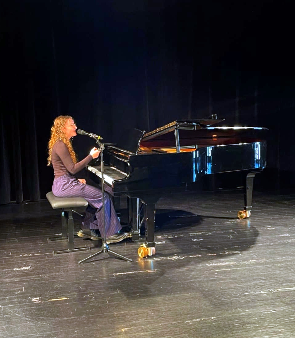 Eine junge Frau sitzt auf einer Bühne an einem schwarzen Flügel und singt in ein Mikrofon. Sie trägt ein dunkelbraunes Oberteil und eine lila Hose. Die Szene ist in warmes Bühnenlicht getaucht, der Hintergrund ist dunkel.