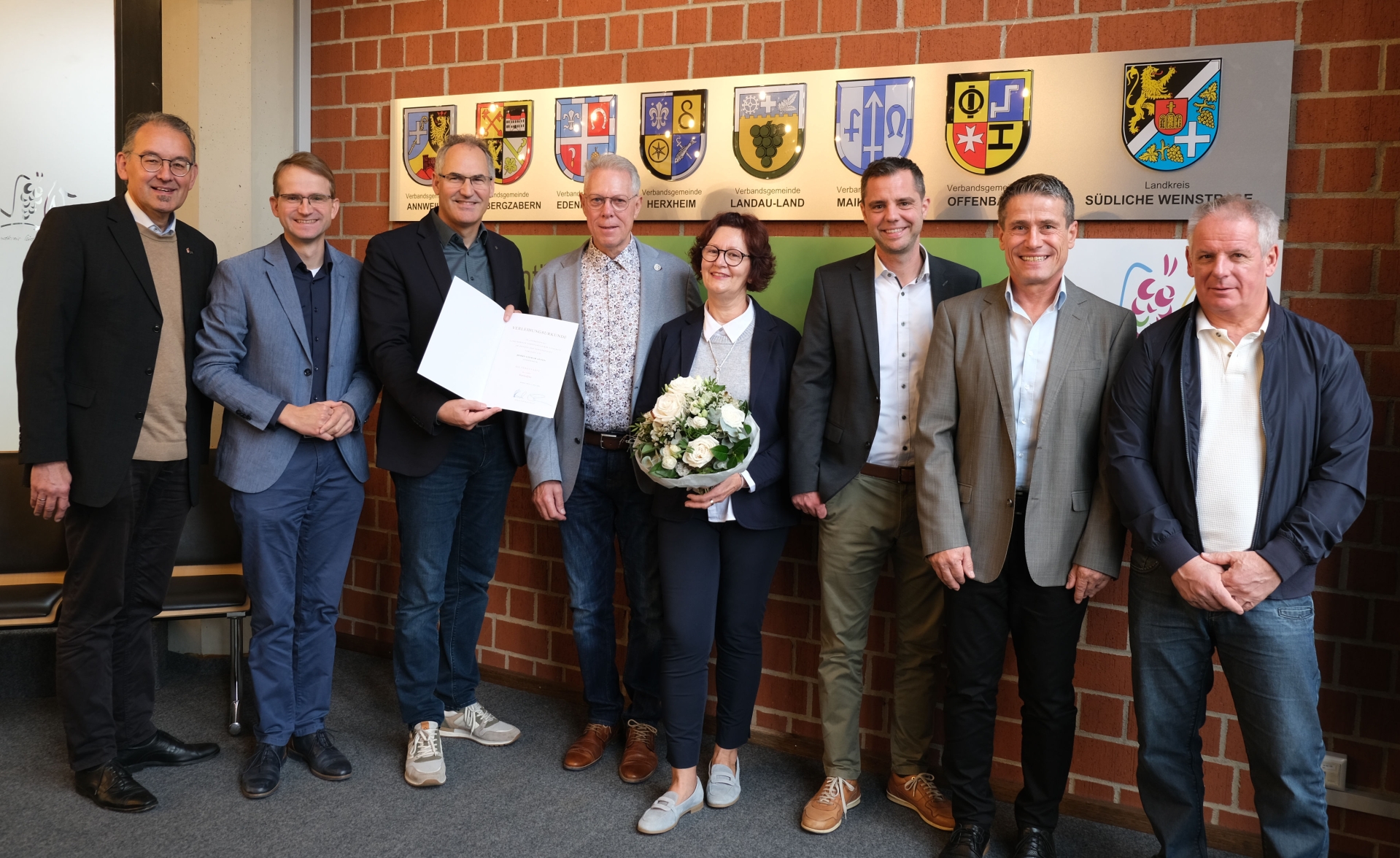 Acht Personen stehen für ein Gruppenbild in einem Raum vor einer Ziegelmauer. In der Mitte der Gruppe hält eine Frau einen Strauß Blumen, ein Mann präsentiert eine Urkunde.