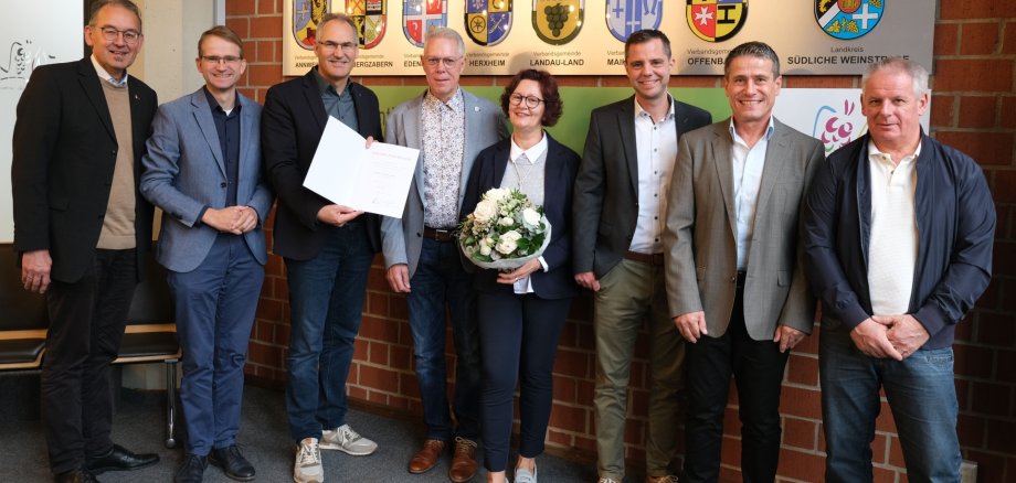 Acht Personen stehen für ein Gruppenbild in einem Raum vor einer Ziegelmauer. In der Mitte der Gruppe hält eine Frau einen Strauß Blumen, ein Mann präsentiert eine Urkunde.