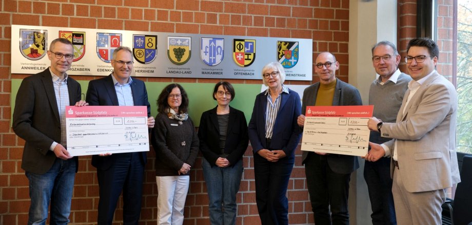 Acht Personen stehen für ein Gruppenbild in einem Raum vor einer Ziegelmauer. Außen halten je zwei Personen zwei große symbolische Schecks.