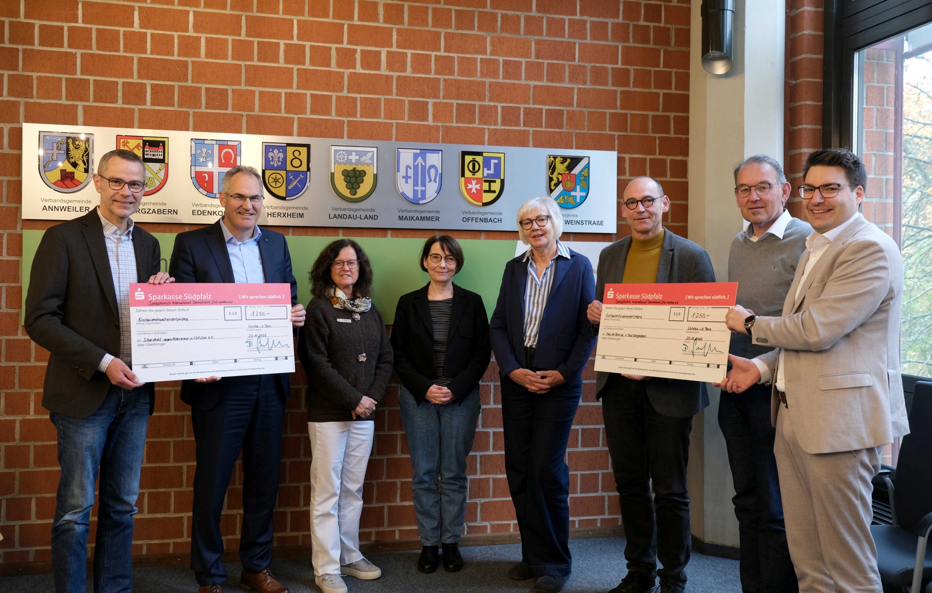 Acht Personen stehen für ein Gruppenbild in einem Raum vor einer Ziegelmauer. Außen halten je zwei Personen zwei große symbolische Schecks.
