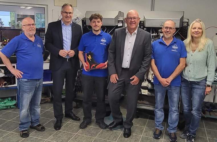 Sechs Personen stehen nebeneinander für ein Gruppenbild und lächeln in die Kamera. Drei Männer tragen jeweils ein blaues Poloshirt mit dem Logo "Wilhelm Orthopädieschuhtechnik & Sanitätsfachhandel". Der junge Mann in der Mitte hält einen schwarz-roten Schuh mit einem gelben Blitz in Händen – sein Meisterstück.