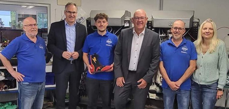 Sechs Personen stehen nebeneinander für ein Gruppenbild und lächeln in die Kamera. Drei Männer tragen jeweils ein blaues Poloshirt mit dem Logo "Wilhelm Orthopädieschuhtechnik & Sanitätsfachhandel". Der junge Mann in der Mitte hält einen schwarz-roten Schuh mit einem gelben Blitz in Händen – sein Meisterstück.