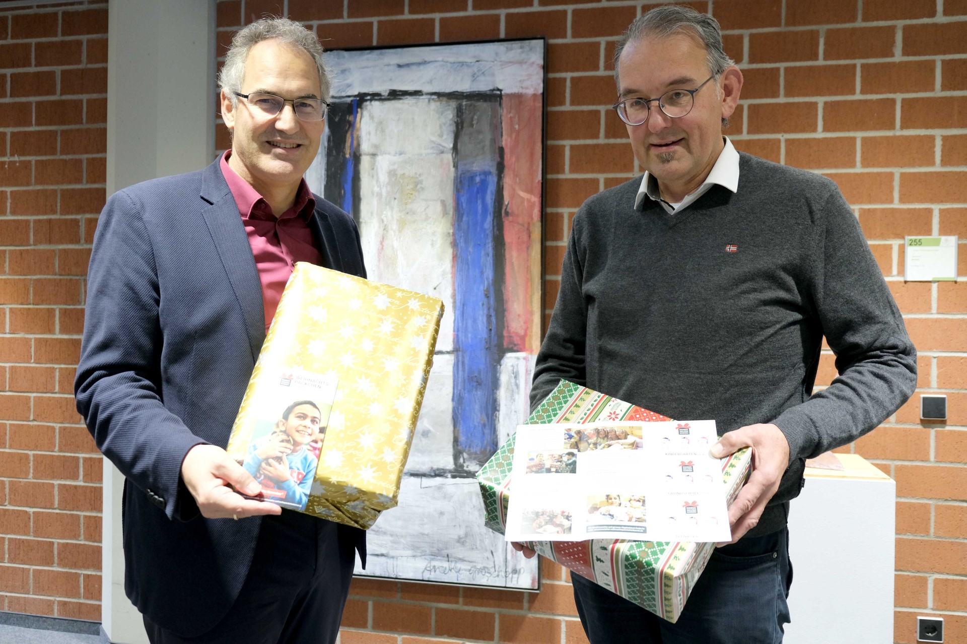 Landrat Dietmar Seefeldt und Erster Kreisbeigeordneter Georg Kern stehen im Kreishaus vor einer Ziegelwand und halten je ein in weihnachtliches Geschenkpapier gehülltes Päckchen in Händen.