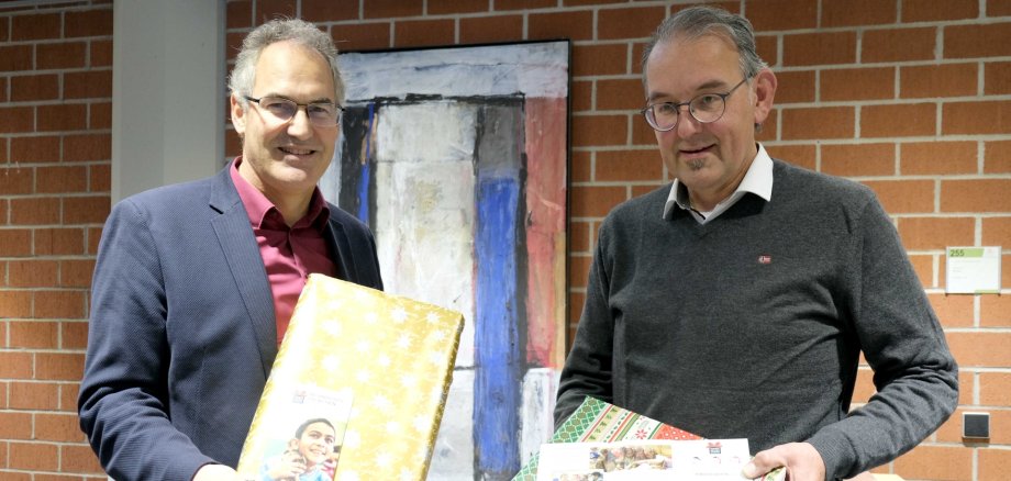 Landrat Dietmar Seefeldt (links) und Erster Kreisbeigeordneter Georg Kern werben für den „Weihnachtspäckchenkonvoi“. Foto: KV SÜW Landrat Dietmar Seefeldt und Erster Kreisbeigeordneter Georg Kern stehen im Kreishaus vor einer Ziegelwand und halten je ein in weihnachtliches Geschenkpapier gehülltes Päckchen in Händen.