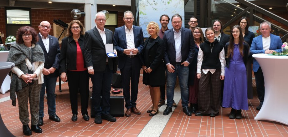 Gruppenfoto im Foyer der Kreisverwaltung Südliche Weinstraße: Mehrere Personen stehen gemeinsam beisammen, in der Mitte wird eine Auszeichnung überreicht. Im Hintergrund sind Kunstwerke und ein Klavier zu sehen.