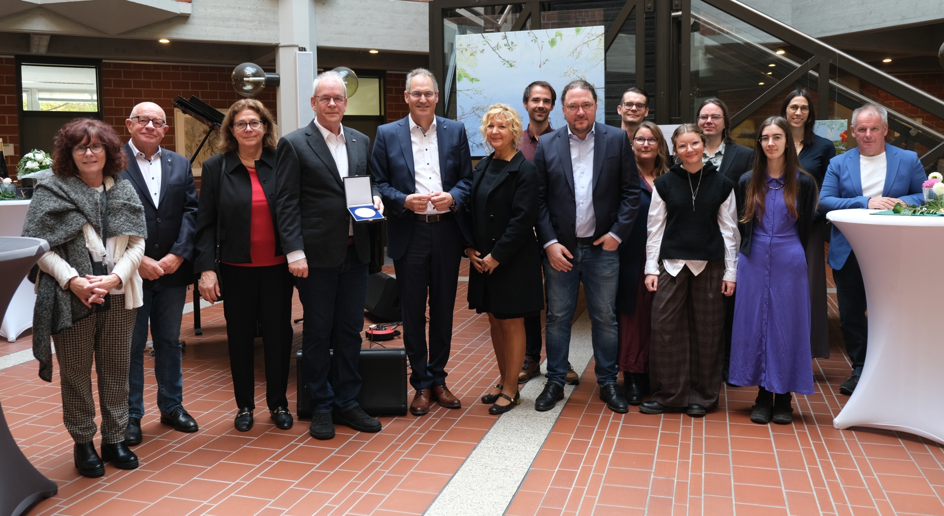 Gruppenfoto im Foyer der Kreisverwaltung Südliche Weinstraße: Mehrere Personen stehen gemeinsam beisammen, in der Mitte wird eine Auszeichnung überreicht. Im Hintergrund sind Kunstwerke und ein Klavier zu sehen.
