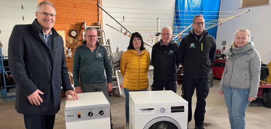 Sechs Personen stehen in einer Reihe in einer Halle hinter zwei Maschinen, einer kleineren alten und einer größeren neuen.