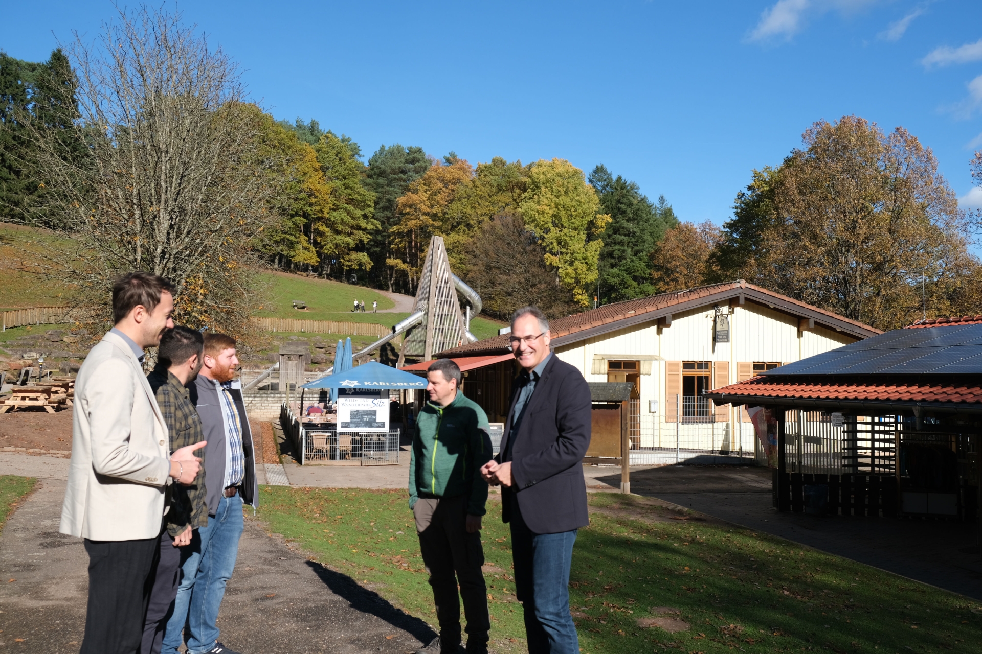 Fünf Männer stehen im Grünen, dahinter zwei Gebäude und eine Riesenrutsche.