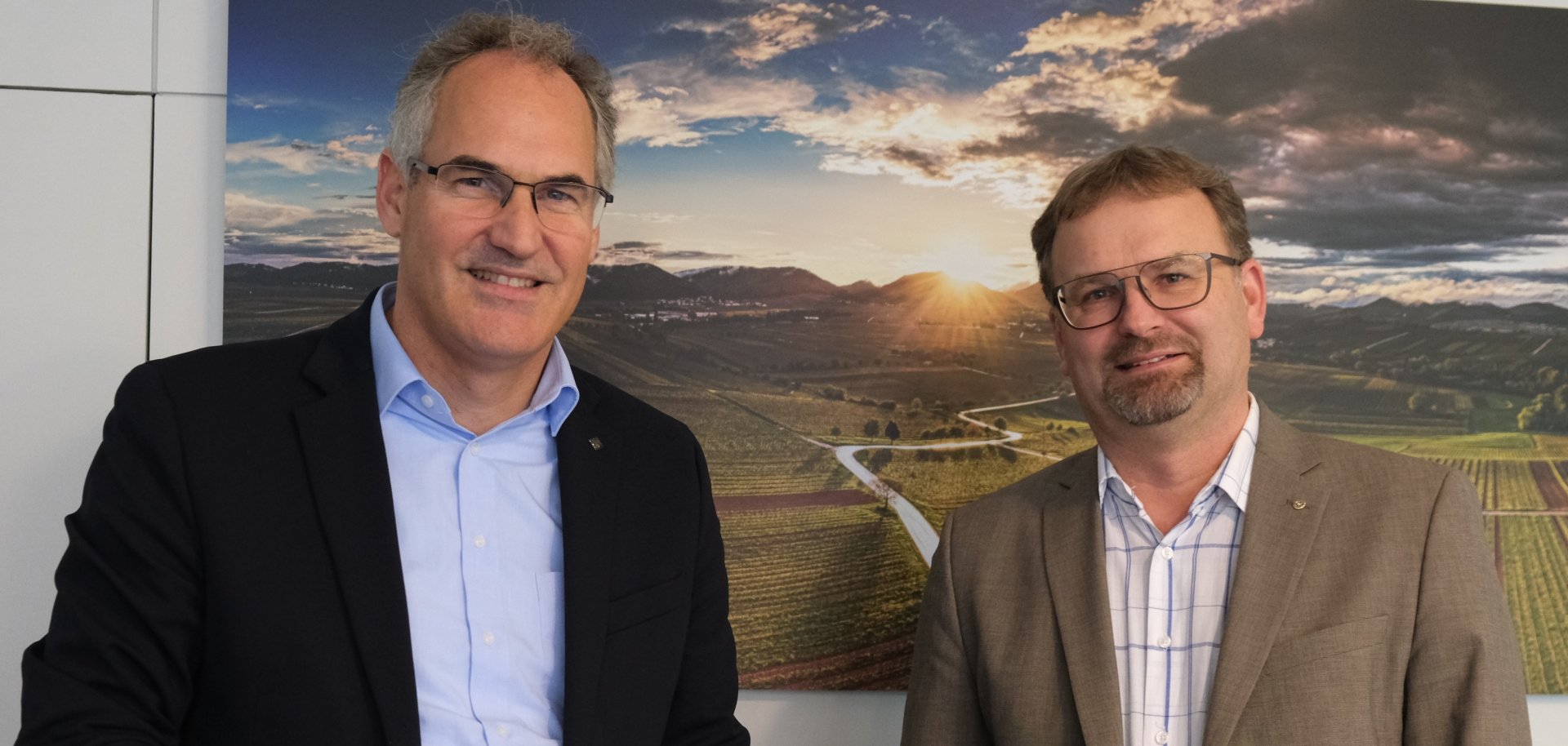 Ansgar Uelhoff und Landrat Dietmar Seefeldt stehen für ein Foto beieinander und lächeln in die Kamera. Im Hintergrund hängt an einer weißen Wand ein Bild einer Weinbergslandschaft im Sonnenuntergang.