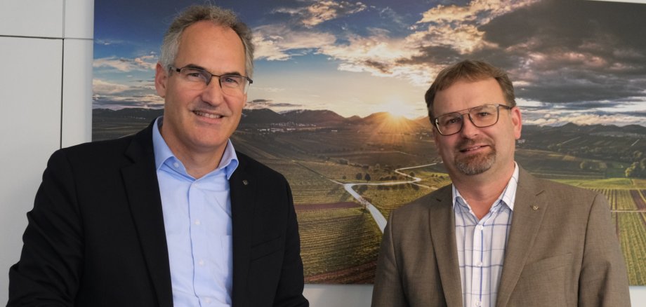 Ansgar Uelhoff und Landrat Dietmar Seefeldt stehen für ein Foto beieinander und lächeln in die Kamera. Im Hintergrund hängt an einer weißen Wand ein Bild einer Weinbergslandschaft im Sonnenuntergang.