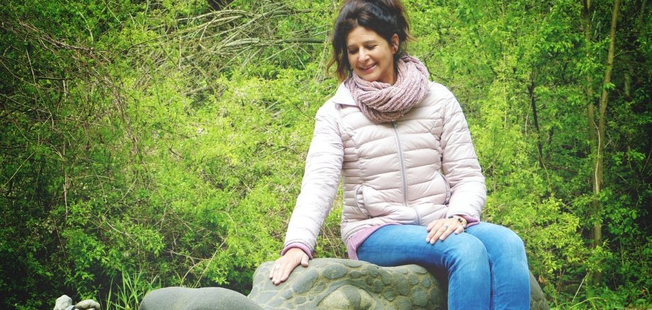 Frau im mittleren Alter sitzt im Freien auf einem steinernen Drachenkopf. Im Hintergrund sind grüne Bäume zu sehen.