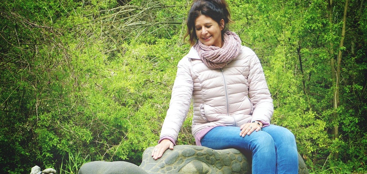 Frau im mittleren Alter sitzt im Freien auf einem steinernen Drachenkopf. Im Hintergrund sind grüne Bäume zu sehen.