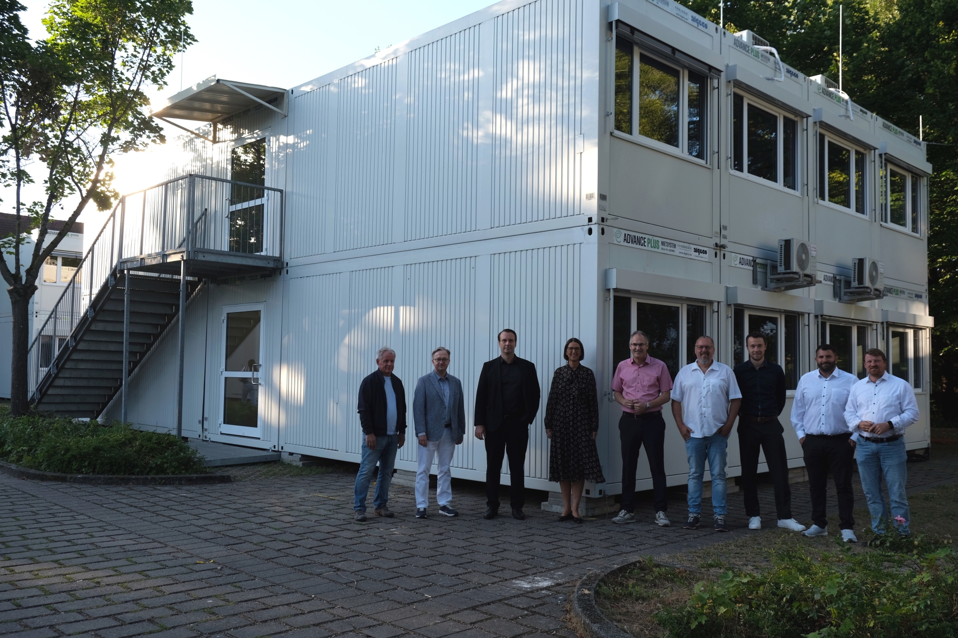 Mehrere Personen stehen vor einem zweigeschossigen weißen Containergebäude mit Außentreppe. Die Gruppe posiert für ein Foto im  Außenbereich des PAMINA-Schulzentrums in Herxheim.