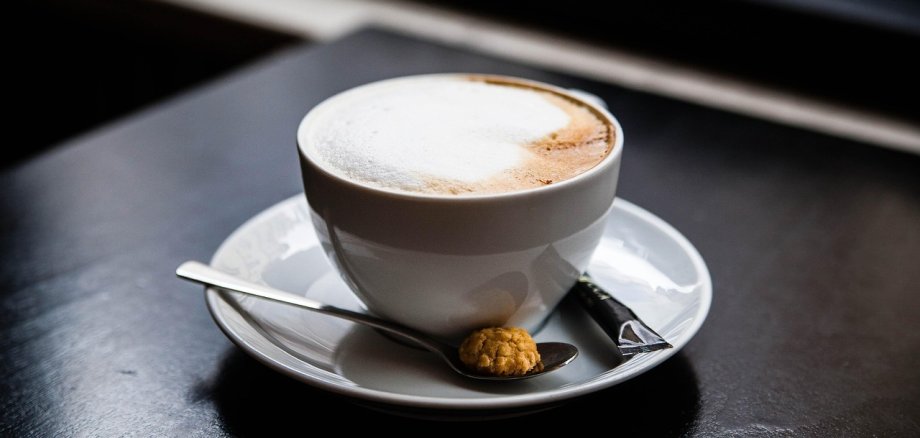 Eine Tasse Cappuccino mit Milchschaum steht auf einem Unterteller. Daneben liegen ein kleiner Löffel mit einem Keks und ein verpacktes Zuckerstäbchen auf einem dunklen Tisch.