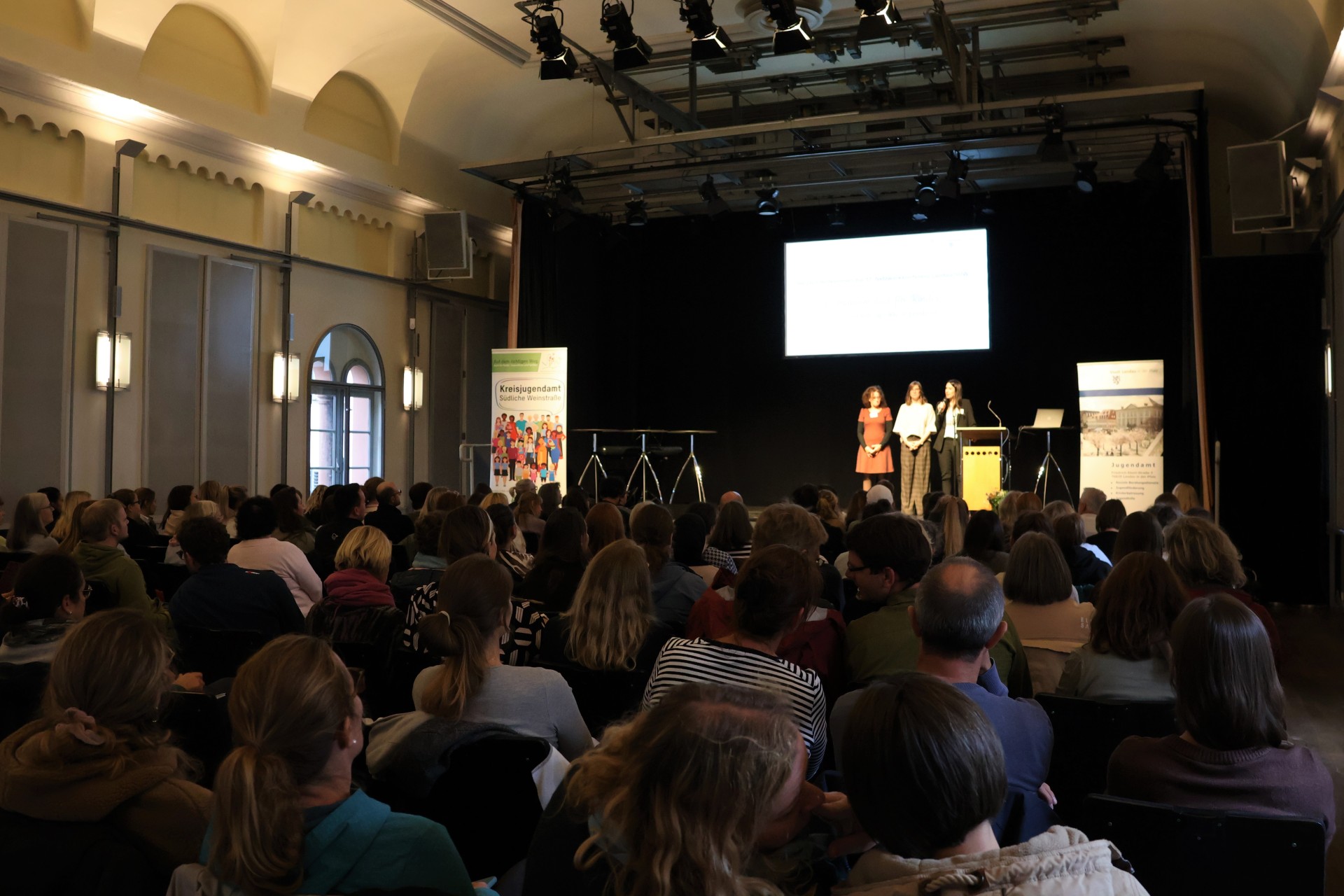 Blick auf eine Bühne in einem Saal mit hoher Decke über das Publikum hinweg. Auf der Bühne stehen drei Personen an beziehungsweise neben einem Rednerpult.