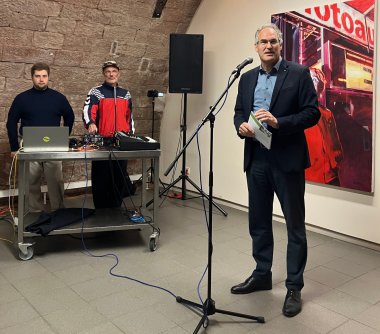 Landrat Dietmar Seefeldt spricht bei der Eröffnung der Kreiskulturtage 2025 im Museum unterm Trifels in Annweiler. Im Hintergrund stehen zwei Musiker an einem DJ-Pult vor einer Sandsteinwand.