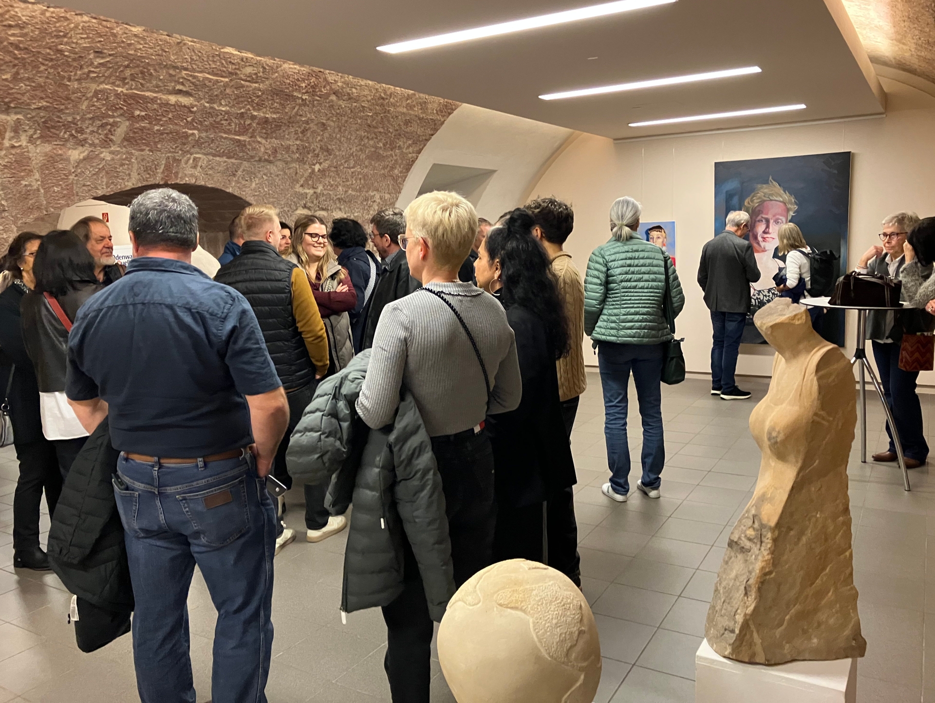 Die Vernissage der Ausstellung von deutschen und französischen Künstlerinnen und Künstlerin im Gewölbekeller des Annweilerer Hohenstaufensaals stieß auf reges Interesse. Im Vordergrund Werke der Bildhauerin Colette Wendel’Ehr, hinten Porträts des Malers Ulli Bomans. Foto: KV SÜW Zahlreiche Besucherinnen und Besucher bei der Vernissage zur Eröffnung der Kreiskulturtage 2025 im Museum unterm Trifels in Annweiler, umgeben von Gemälden und Skulpturen.