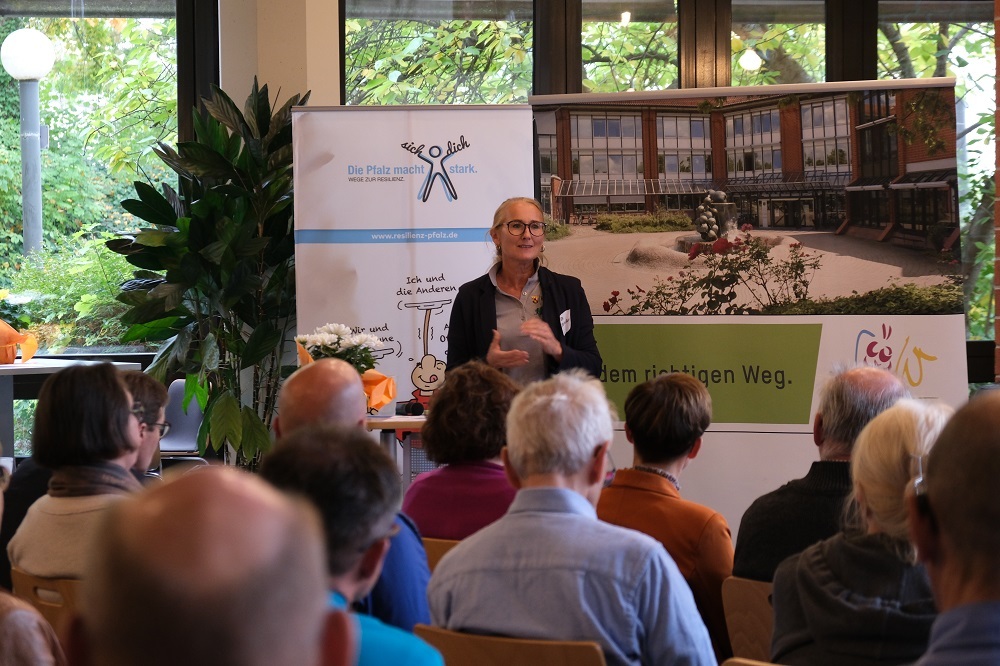 Blick über über Zuschauer auf Stühlen hinweg auf eine Frau, die vor dem Publikum steht und vorzutragen scheint. Hinter ihr sind zwei Rollups zu sehen. Eines zeigt die Außenansicht des Kreishauses in Landau, darunter das Logo der Kreisverwaltung SÜW sowie der Claim "Auf dem richtigen Weg". Das andere ist mit dem Slogan "Die Pfalz macht stark." überschrieben.