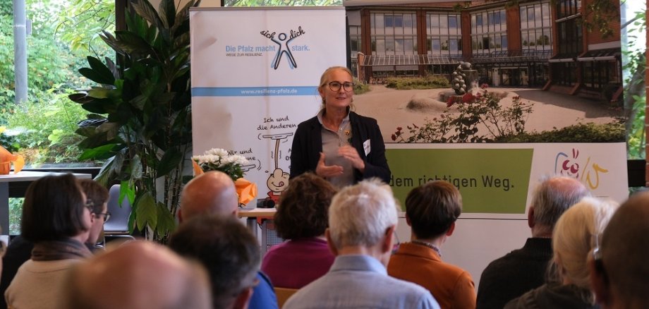 Blick über über Zuschauer auf Stühlen hinweg auf eine Frau, die vor dem Publikum steht und vorzutragen scheint. Hinter ihr sind zwei Rollups zu sehen. Eines zeigt die Außenansicht des Kreishauses in Landau, darunter das Logo der Kreisverwaltung SÜW sowie der Claim "Auf dem richtigen Weg". Das andere ist mit dem Slogan "Die Pfalz macht stark." überschrieben.