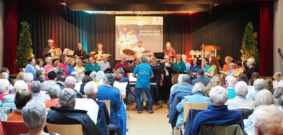 Blick durch einen Mittelgang zwischen vollbesetzen Stuhlreihen zu einer Bühne, auf der mehrere Musiker spielen. Vor der Bühne steht ein gemischter Chor. Auf einem Banner auf der Bühne ist die Aufschrift "Gemeinsam in ganz Deutschland singen" zu lesen.